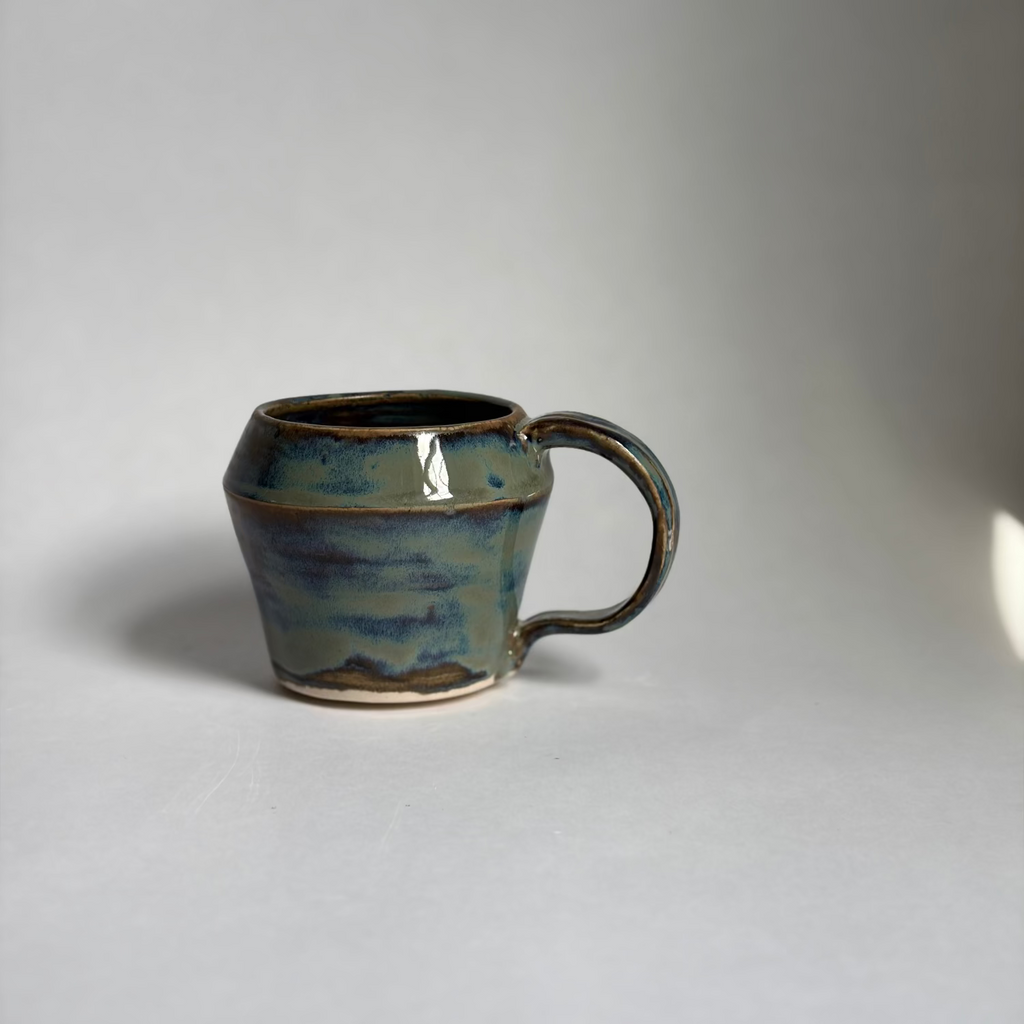 A handmade ceramic mug with a textured, tapered shape and a curved handle, featuring a glossy glaze with blue, teal, and brown earthy tones, set against a plain light gray background.