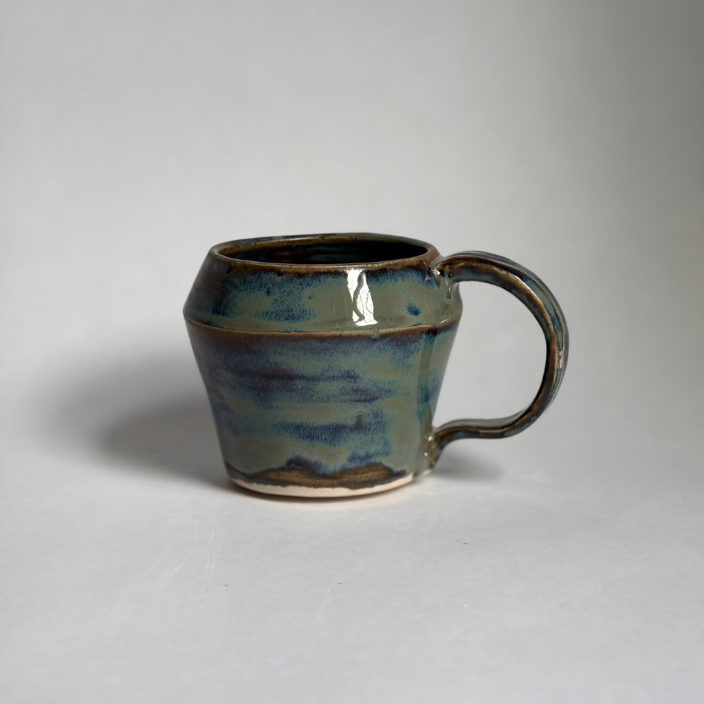 A handmade ceramic mug with a rounded body and a looped handle, featuring a glossy glaze with blue, green, and brown earthy tones, set against a plain light gray background.
