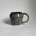 A handmade ceramic mug with a rounded body and a looped handle, featuring a glossy glaze with blue, green, and brown earthy tones, set against a plain light gray background.