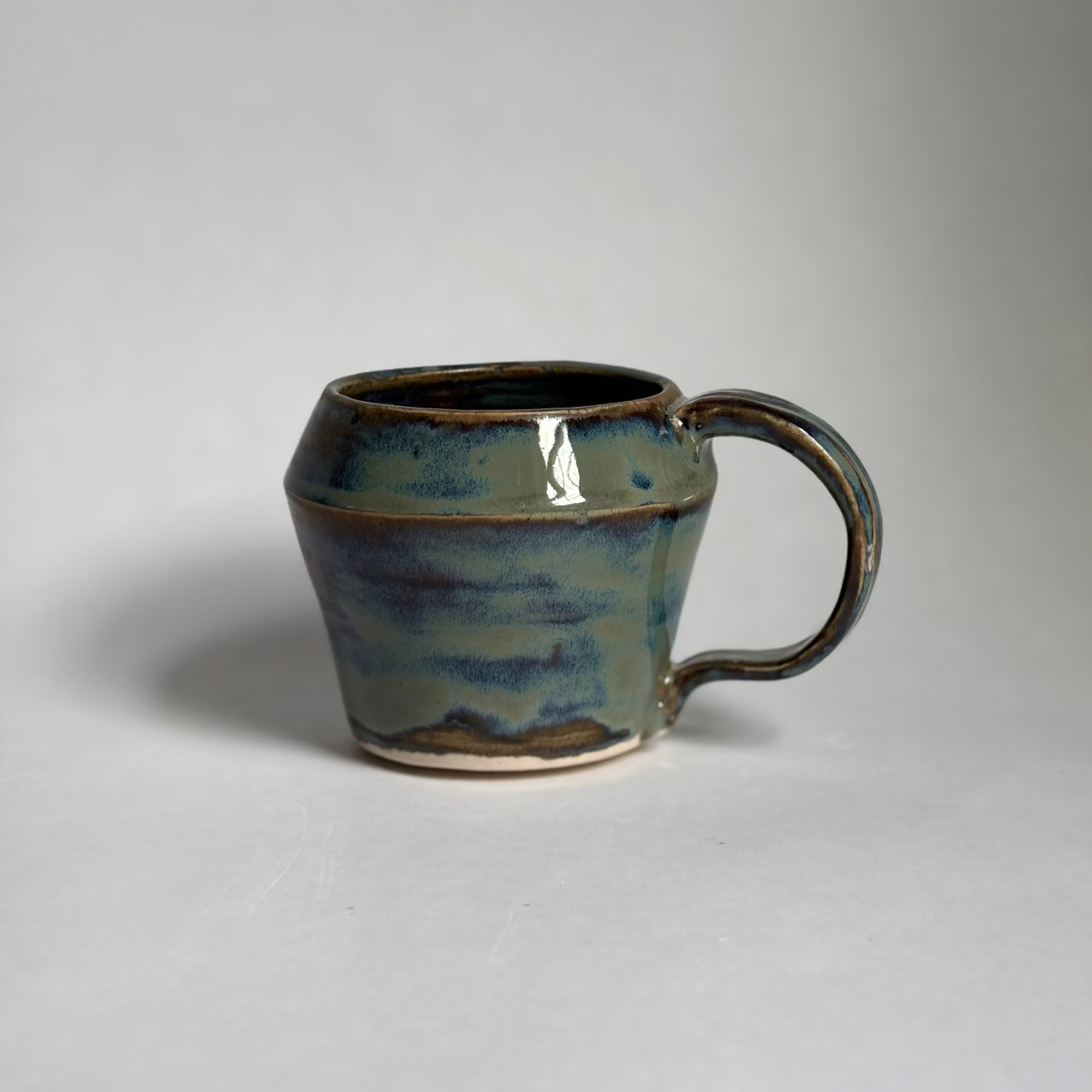 A handmade ceramic mug with a rounded body and a looped handle, featuring a glossy glaze with blue, green, and brown earthy tones, set against a plain light gray background.