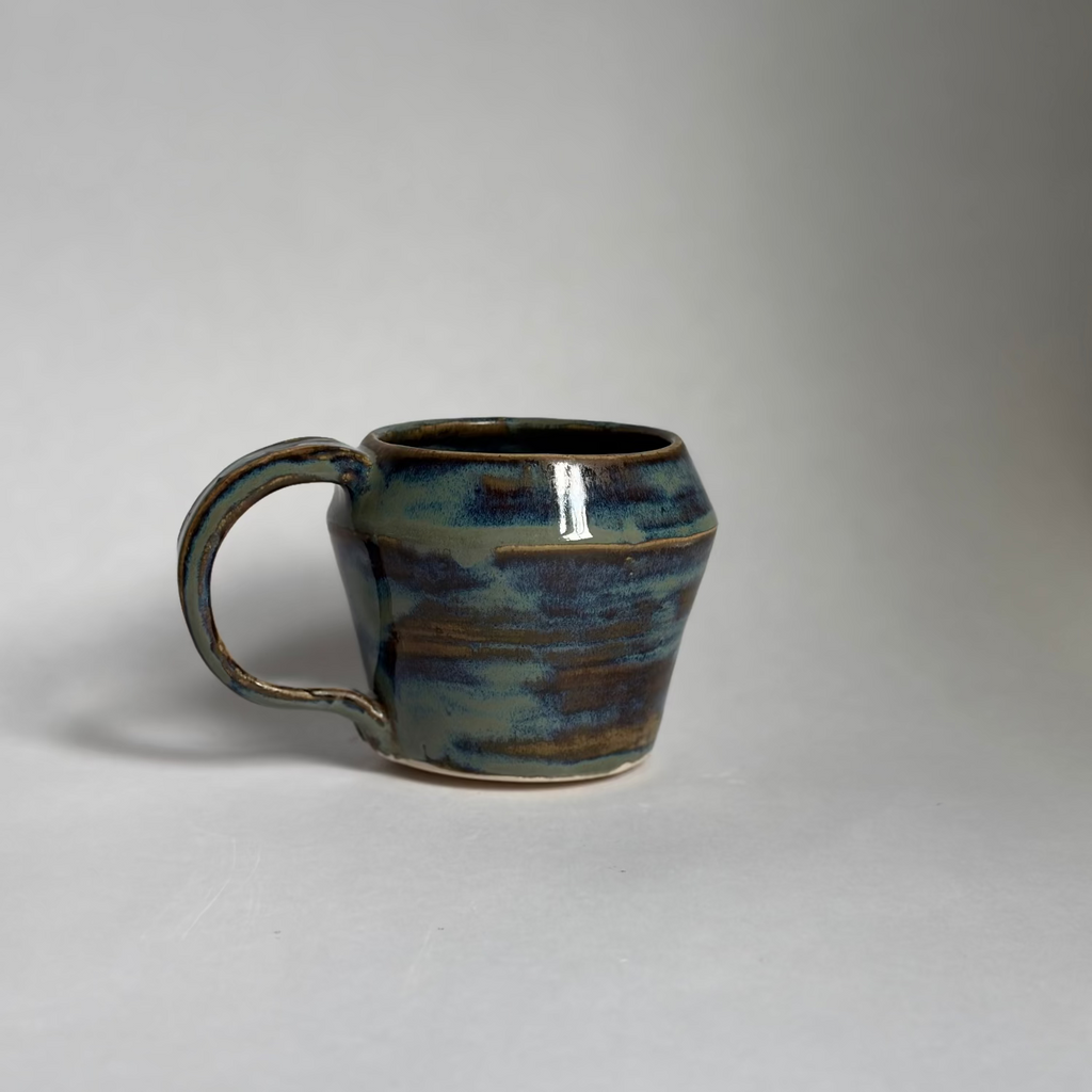A handmade ceramic mug with a rounded body and a looped handle, featuring a glossy glaze with blue, green, and brown earthy tones, set against a plain light gray background.