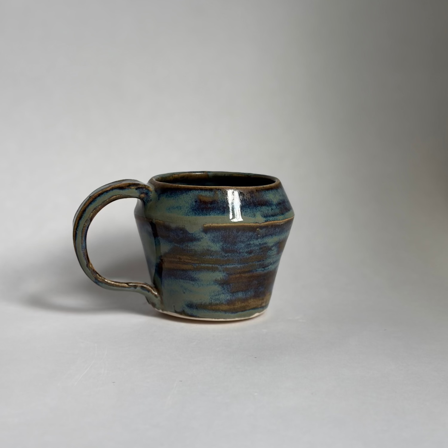A handmade ceramic mug with a rounded body and a looped handle, featuring a glossy glaze with blue, green, and brown earthy tones, set against a plain light gray background.