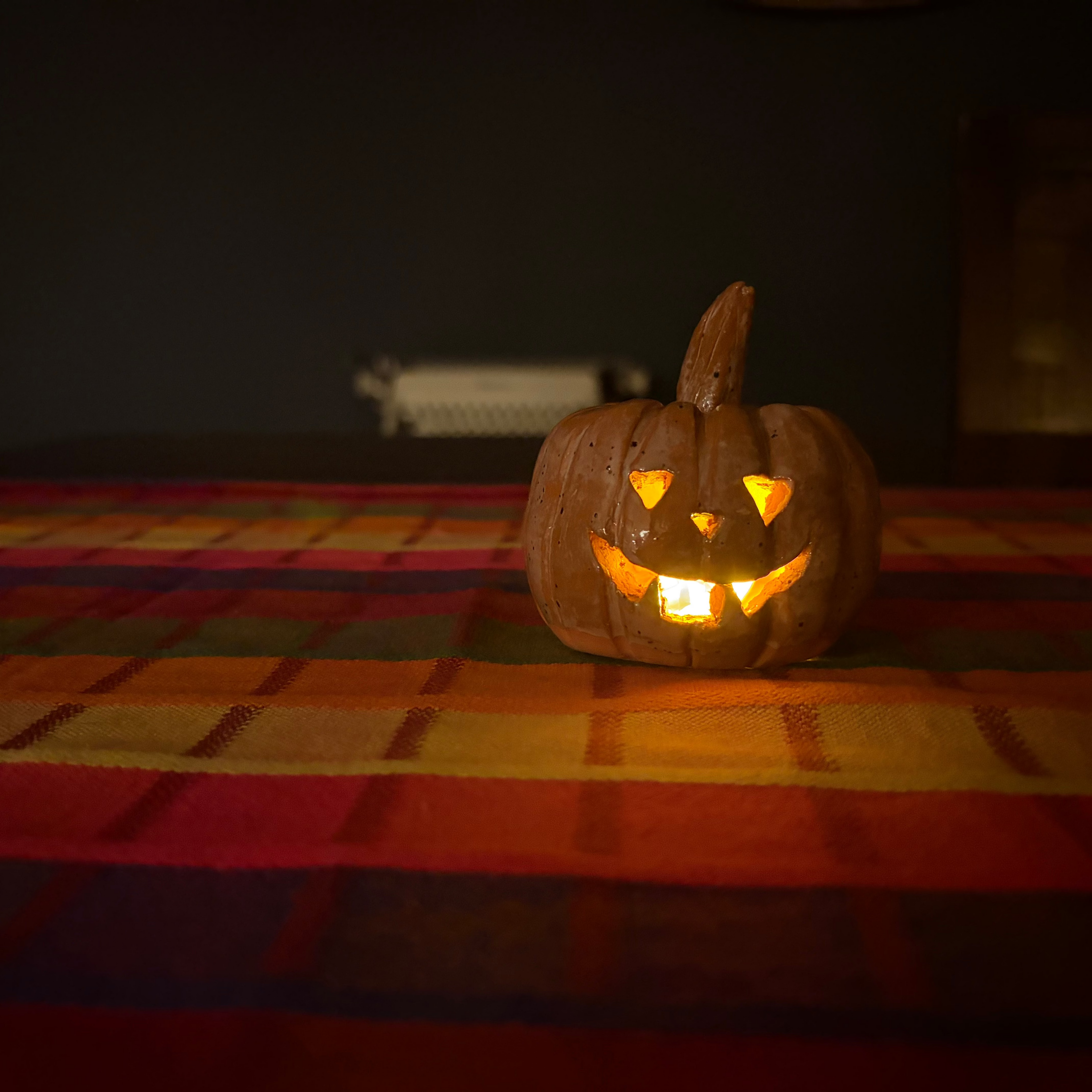 A carved jack-o'-lantern pumpkin with a lit orange glow inside, placed on a striped red, orange, and yellow blanket against a dark background.