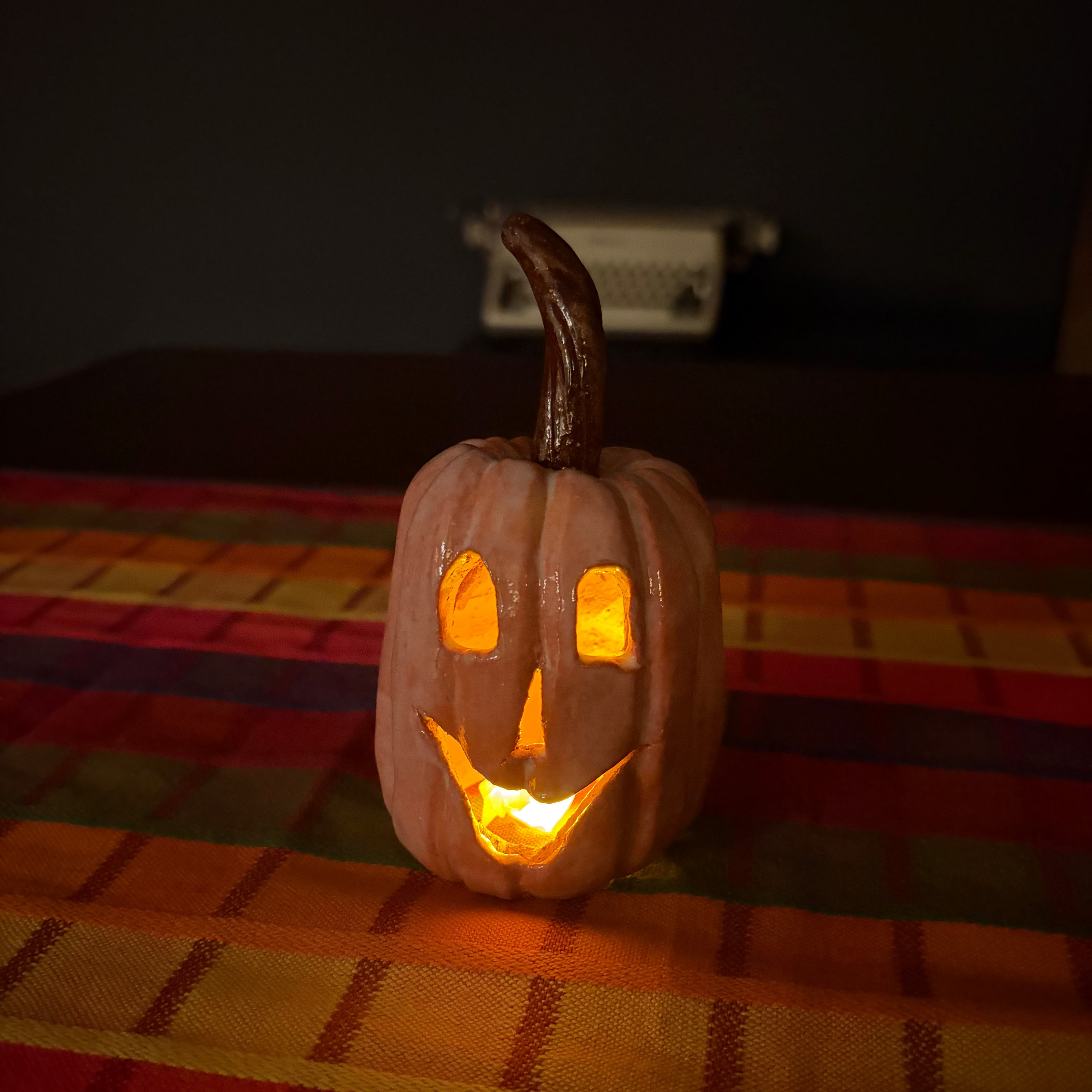 A carved jack-o'-lantern pumpkin with a lit orange glow inside, placed on a red, yellow, and green plaid blanket in a dimly lit room.