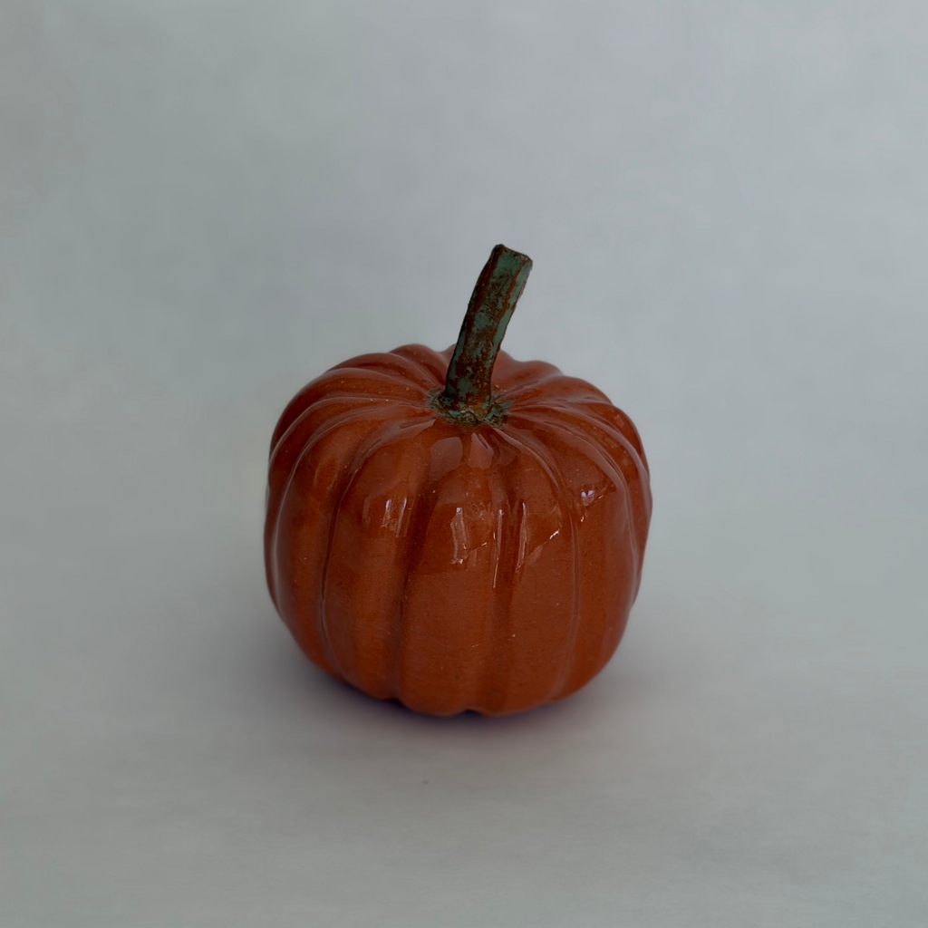 Single orange ceramic pumpkin with a green-tinged stem, photographed on a neutral white background
