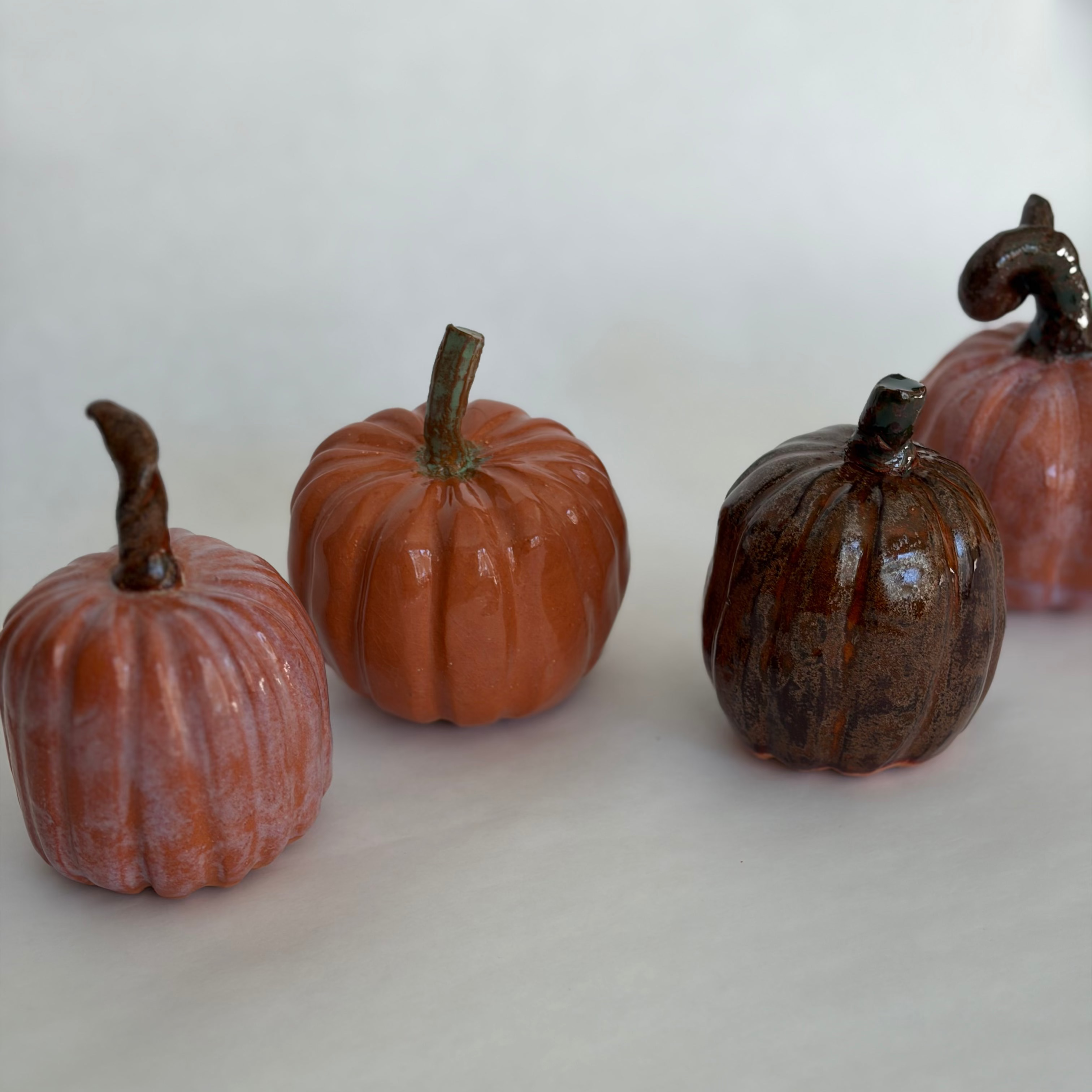 Four ceramic pumpkins in varying earthy glazes — blush pink, burnt orange, and dark brown — arranged in a loose cluster on a white surface
