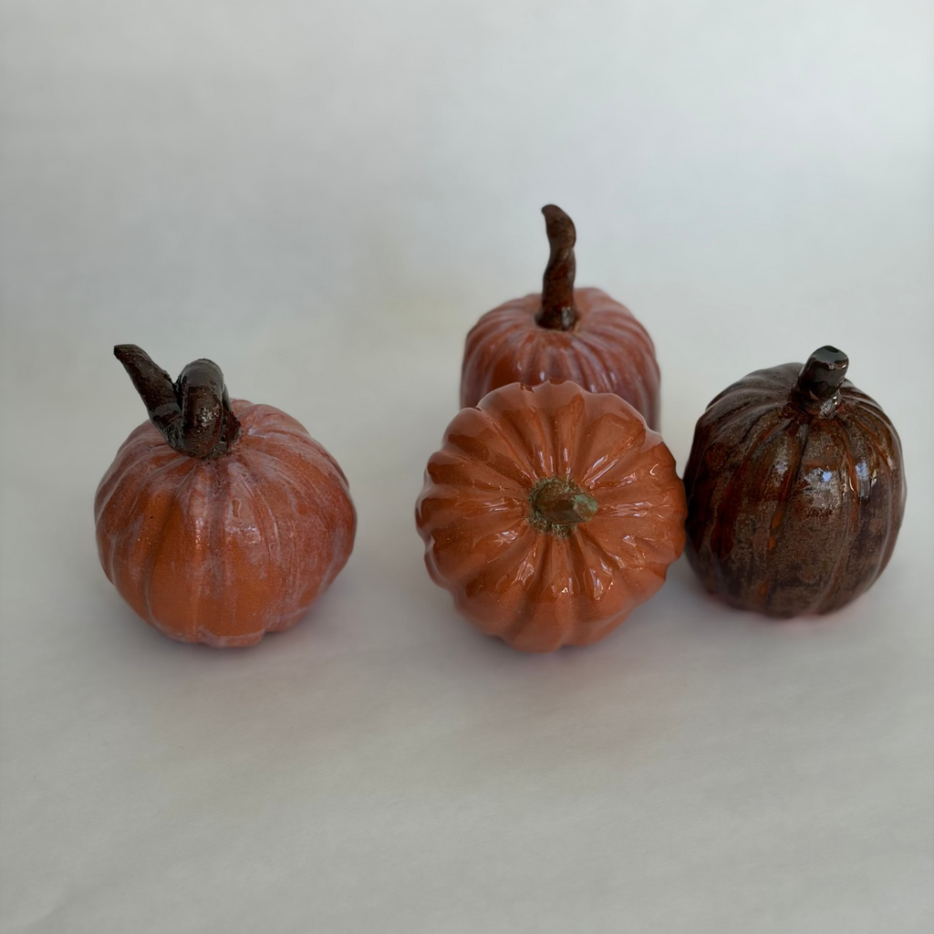 Group of four ceramic pumpkins in terracotta, blush, and deep brown glazes, photographed from above on a white surface
