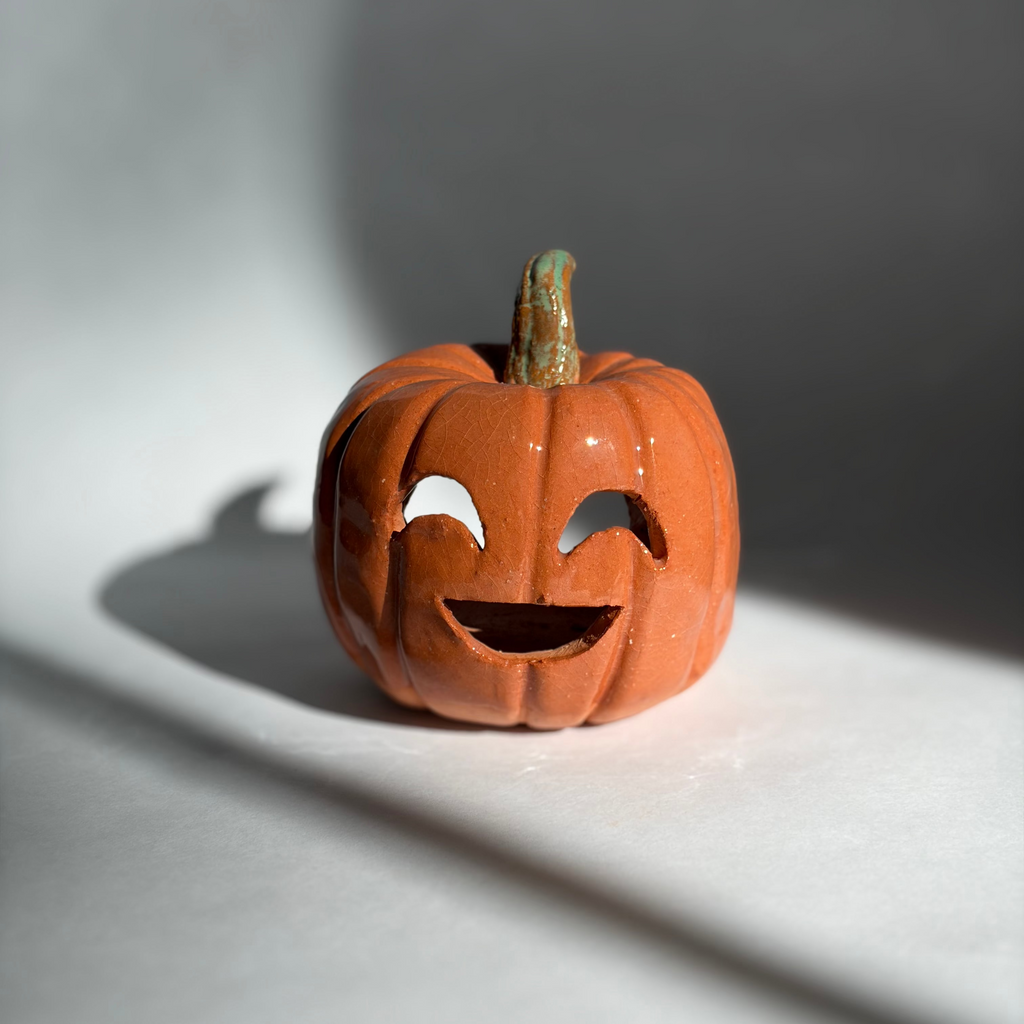 A small carved orange jack-o'-lantern pumpkin figurine with a green stem, photographed on a white surface with dramatic window-light shadows cast across the scene.