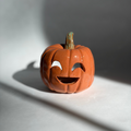 A small carved orange jack-o'-lantern pumpkin figurine with a green stem, photographed on a white surface with dramatic window-light shadows cast across the scene.