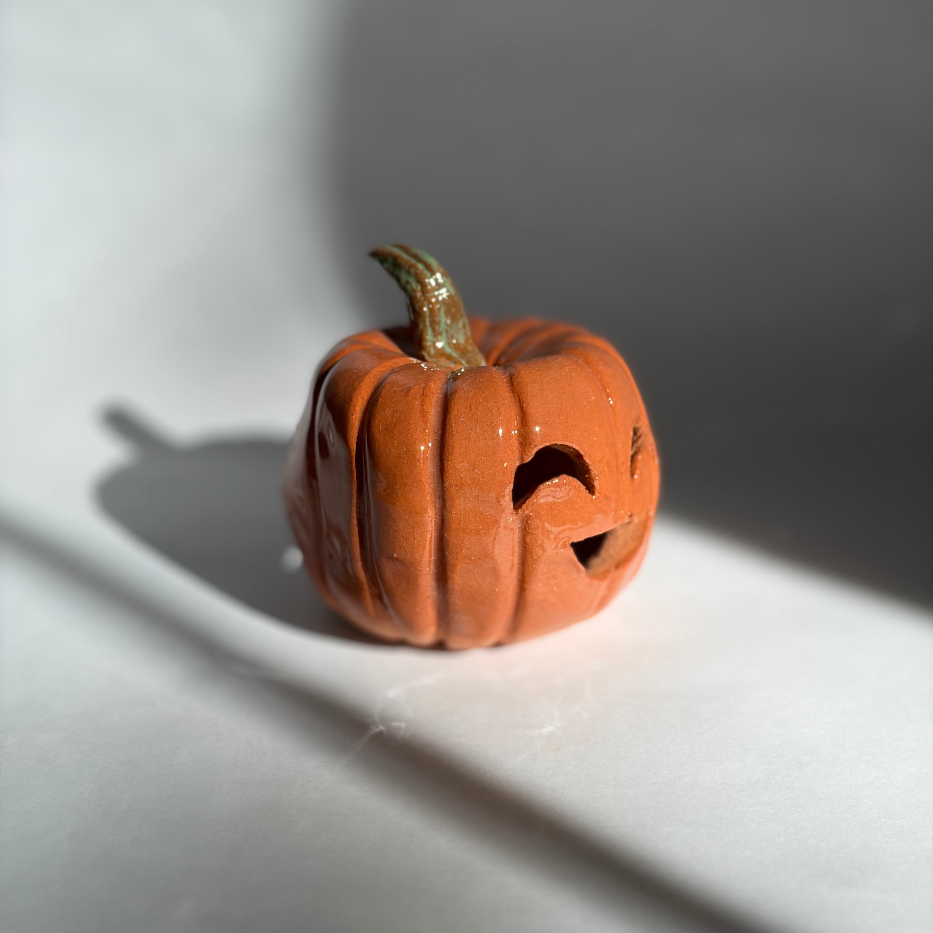 A small carved orange jack-o'-lantern pumpkin figurine with a green stem, photographed on a white surface with dramatic window-light shadows cast across the scene.