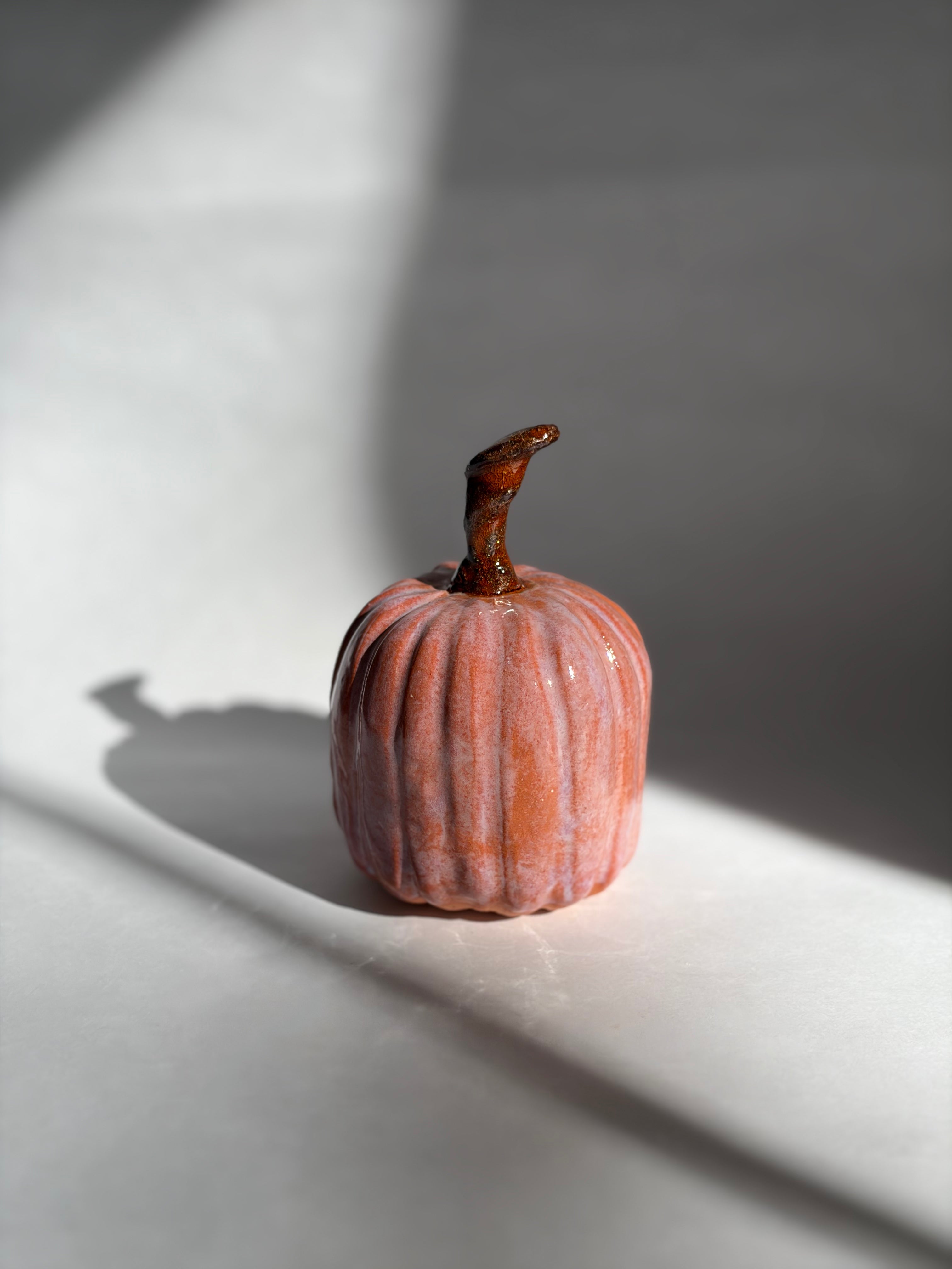 Single blush-pink ceramic pumpkin with a long curved stem, photographed in warm directional light on a white ledge
