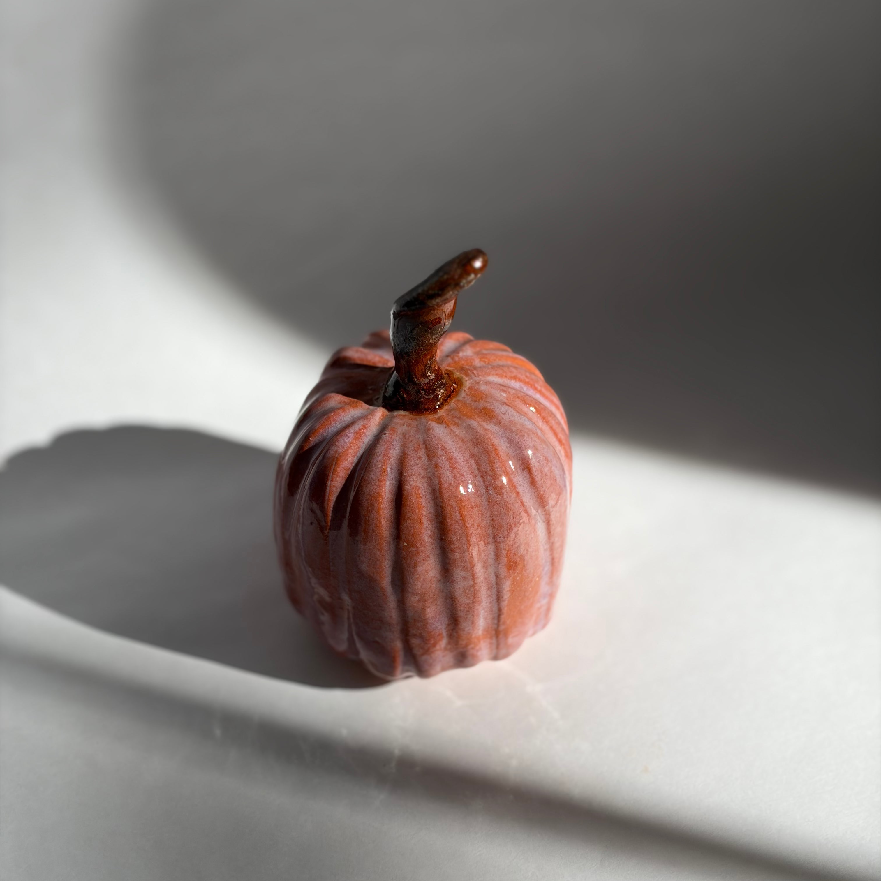 Single terracotta-blush ceramic pumpkin with a dark curved stem, lit with dramatic side light on a white surface
