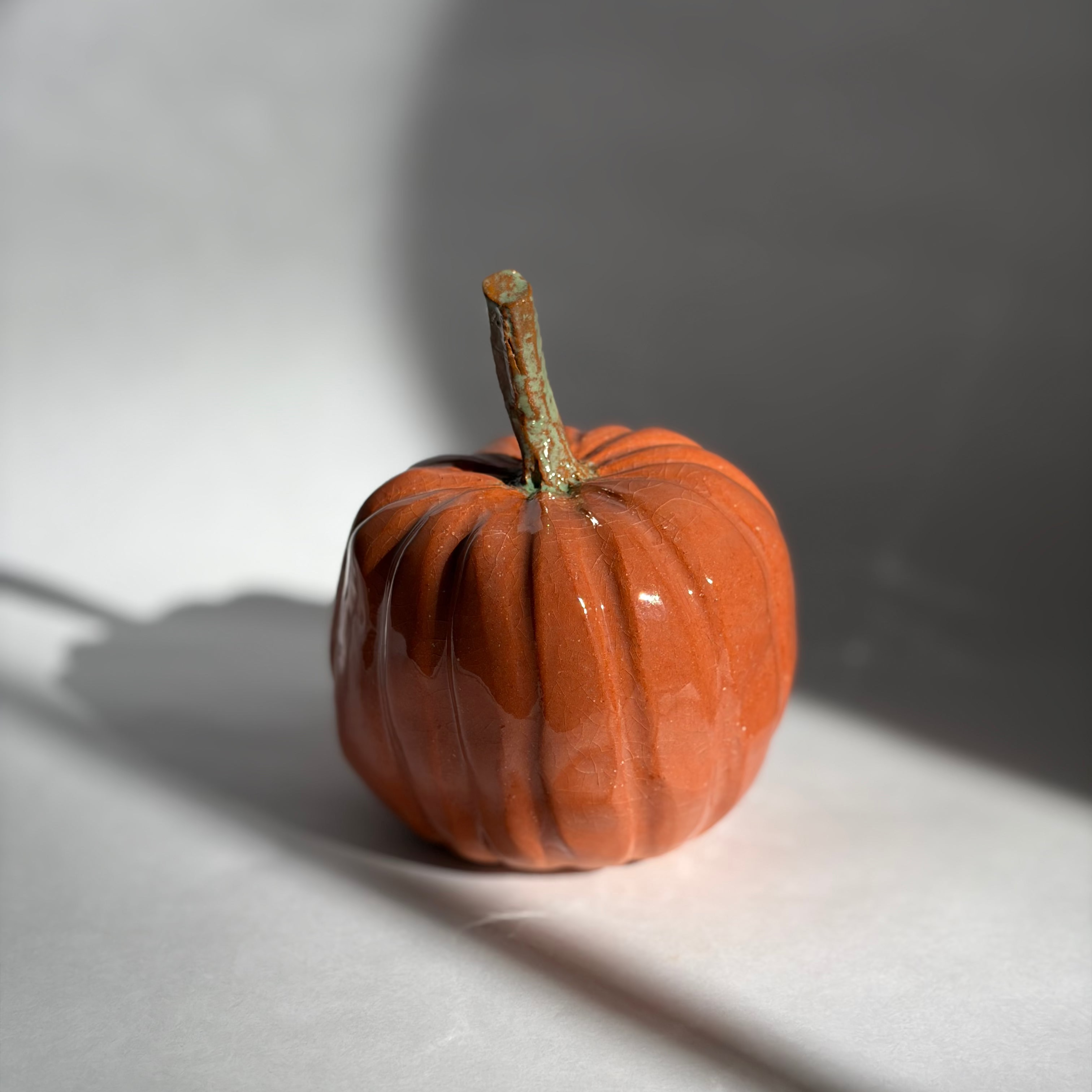 Single deep orange ceramic pumpkin with a straight green-brown stem, photographed in warm directional light with a soft shadow
