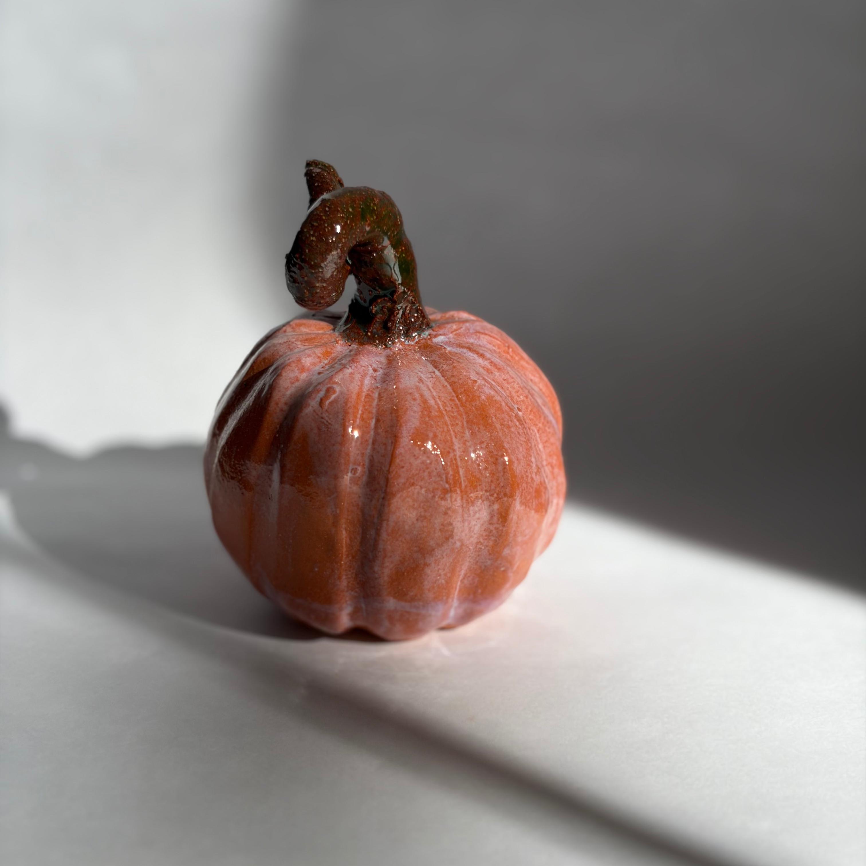 Single blush-terracotta ceramic pumpkin with a twisted dark stem, shown in soft side lighting on a white surface
