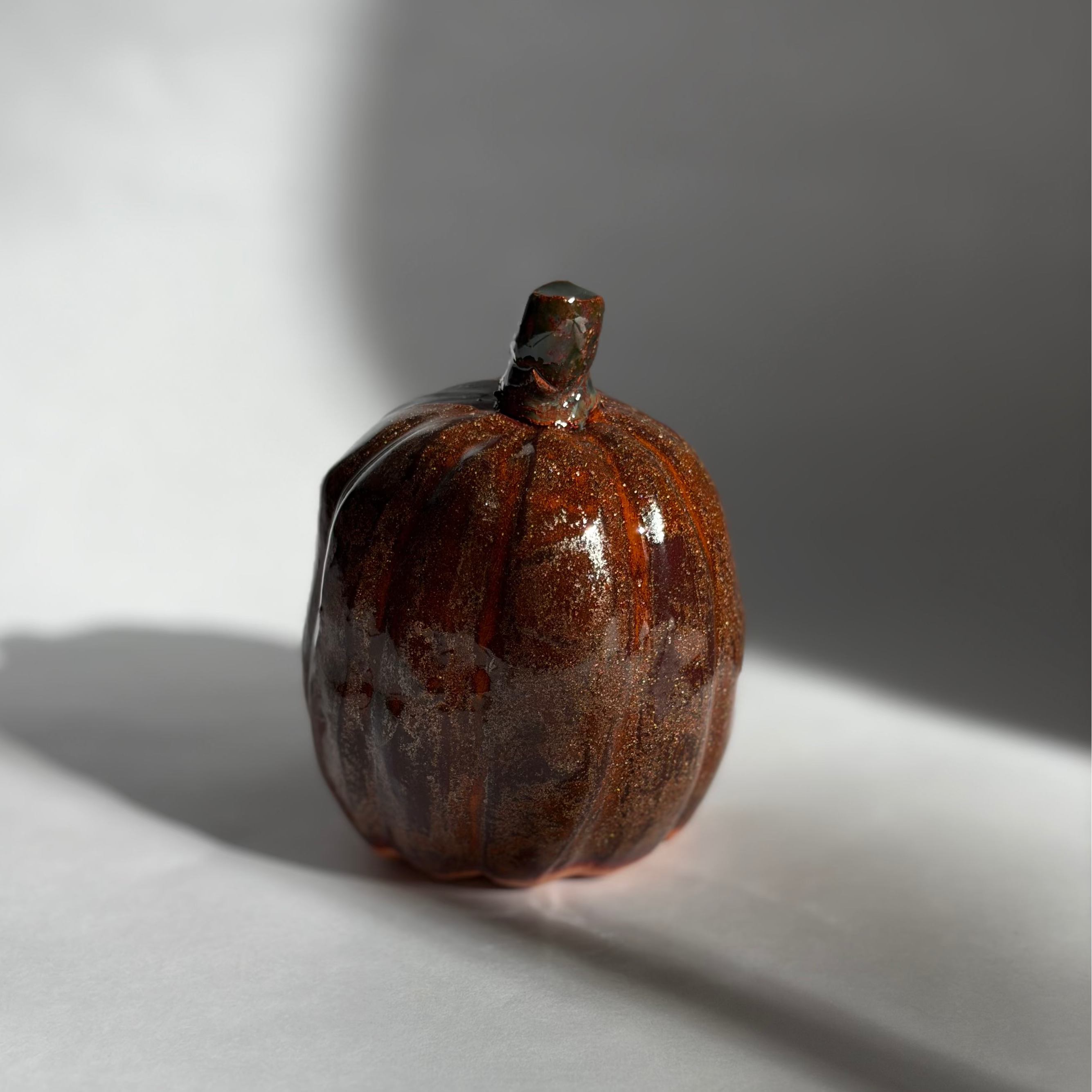 Single deep brown ceramic pumpkin with a short dark stem and rich, speckled glaze, lit with warm directional light
